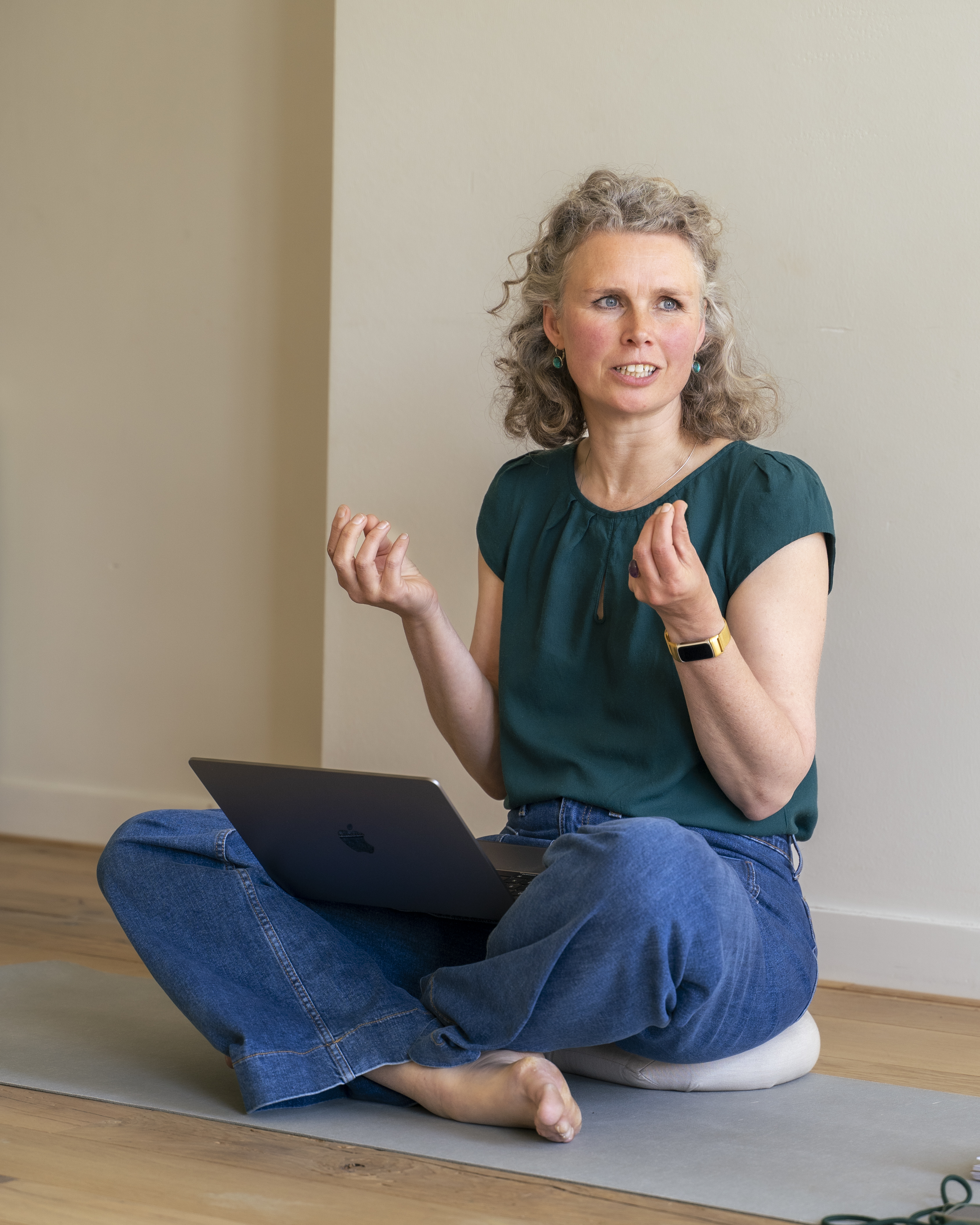 Woman sitting cross legged on a yoga mat with a laptop on her lap and explaining an exercise.