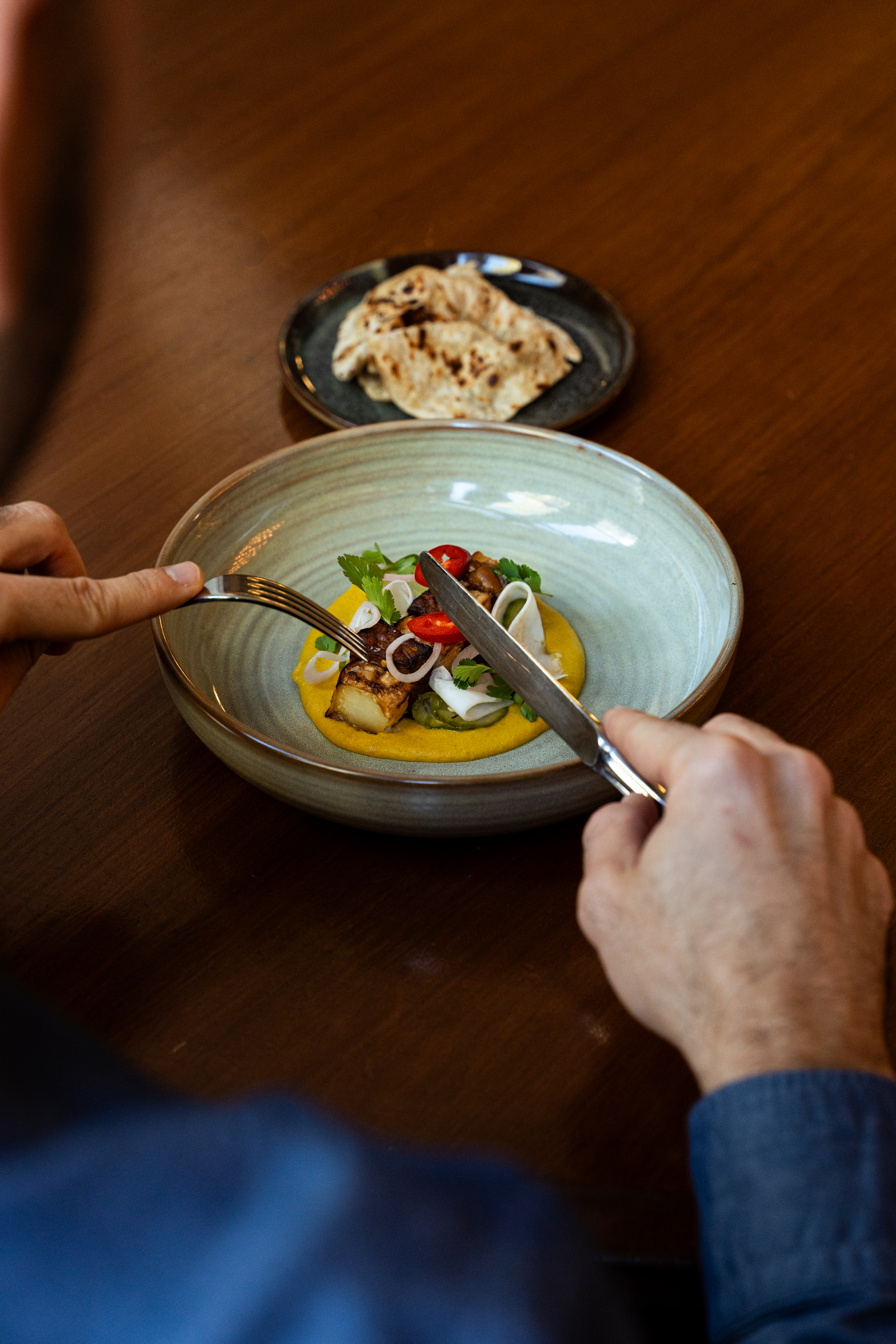 Over the shoulder of a man holding cutlery and eating a dish at a restaurant.
