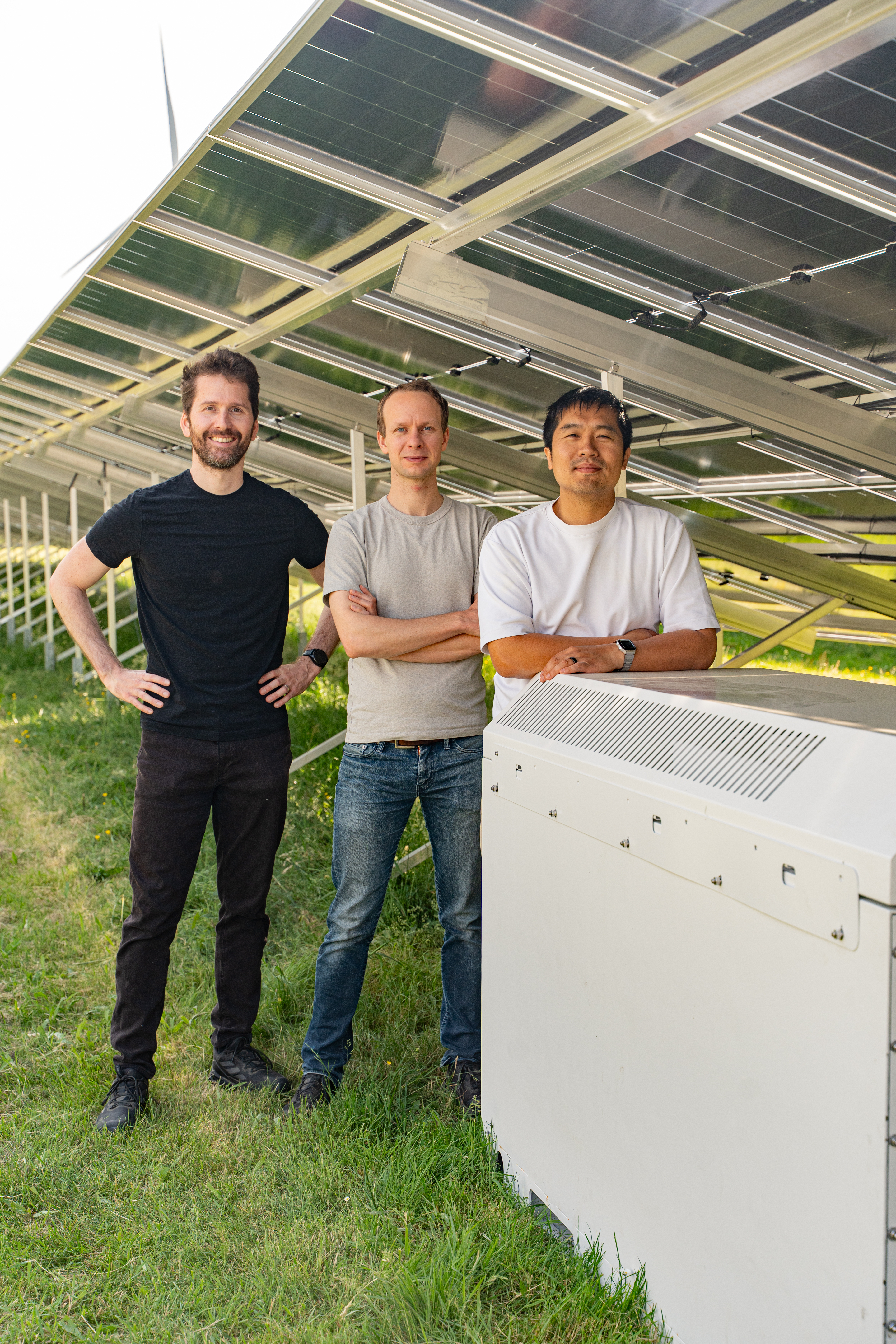 Three men standing under solar panels next to the battery for the panels which they developed.