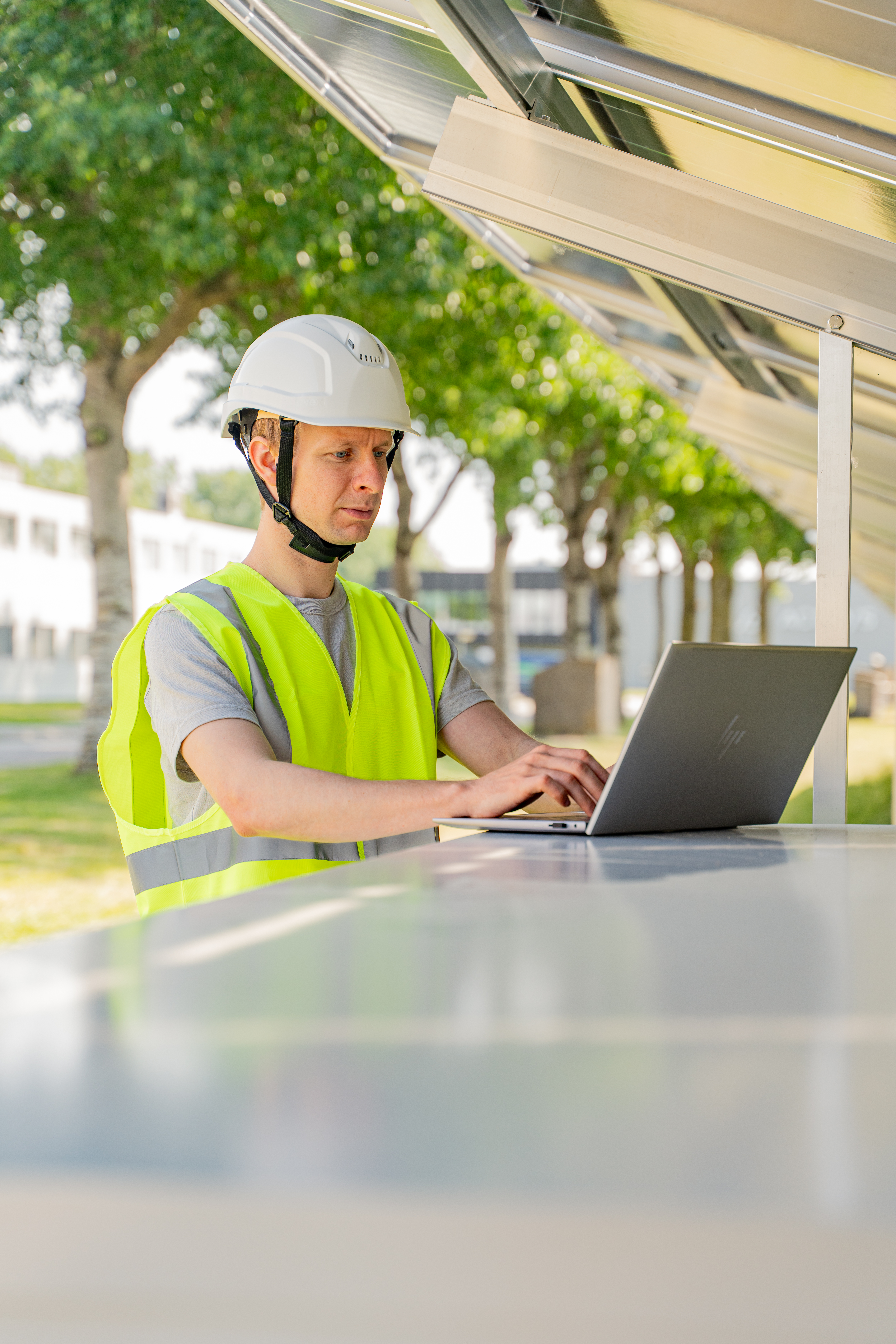 Man wearing fluorescent vest and a white safety helmet standing outside working on a laptop.